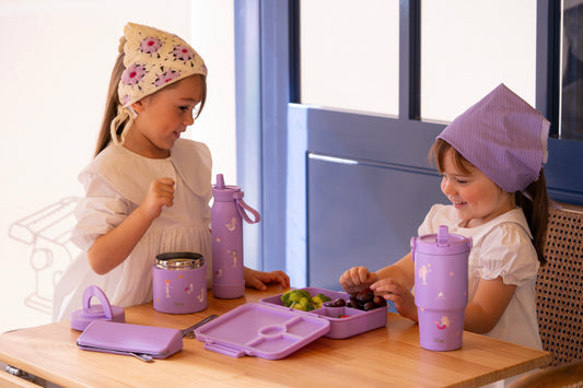 Two girls at a table with purple compartment lunch box, food jar, and water bottle
